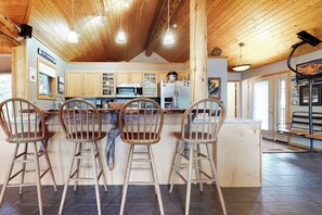 A kitchen with wooden cabinets, a central island with bar stools, and a ceiling with exposed beams. - Dog-Friendly Home with Free WiFi, Private Hot Tub, and Great Views - Near Golfing (Big Sky)