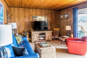 Ein Zimmer mit Holzwänden, einem Fernseher, blauen und roten Sofas, einem kleinen Tisch mit Zeitschriften und Blick auf eine verschneite Landschaft.