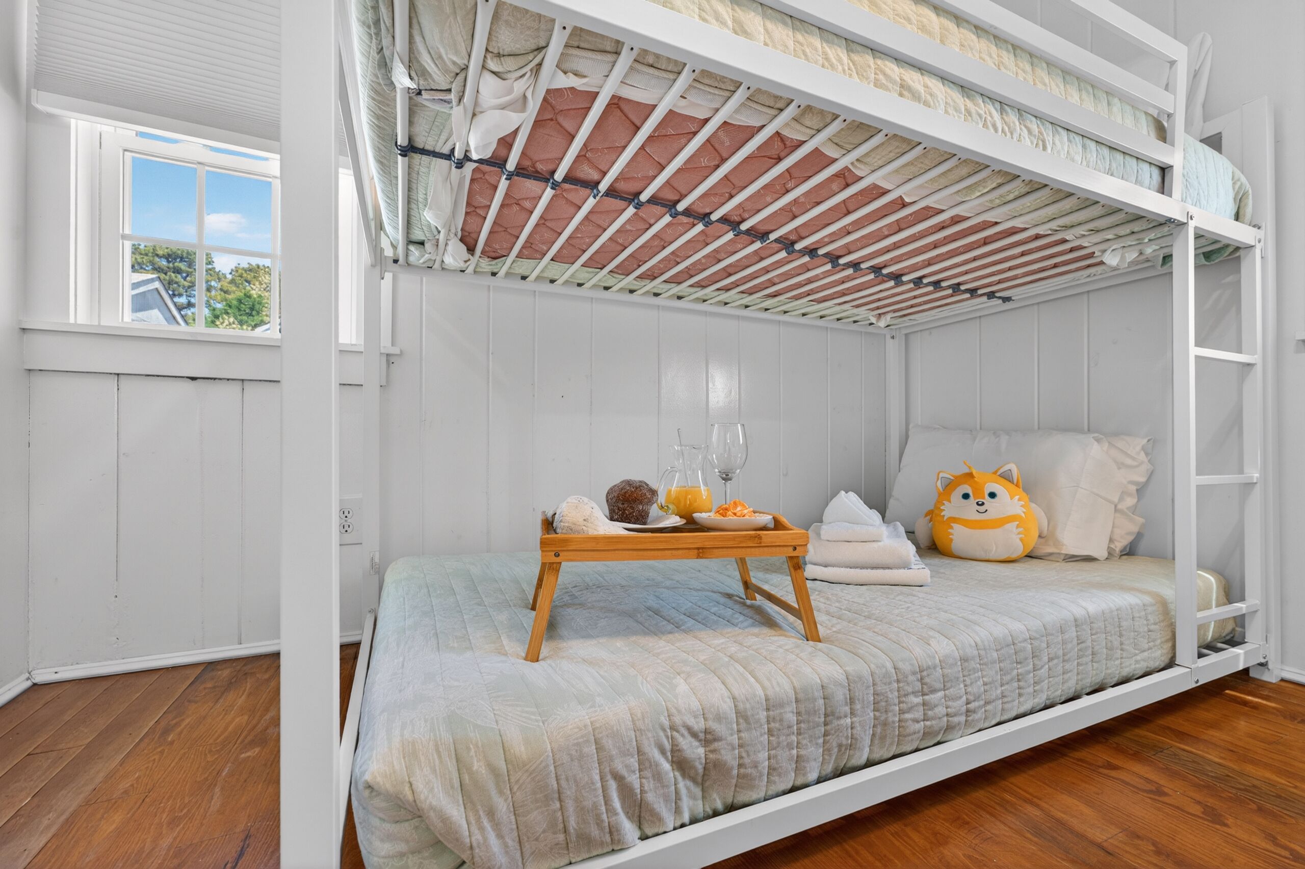 A bunk bed room with a wooden tray table, a plush toy, and a window with a view of trees.