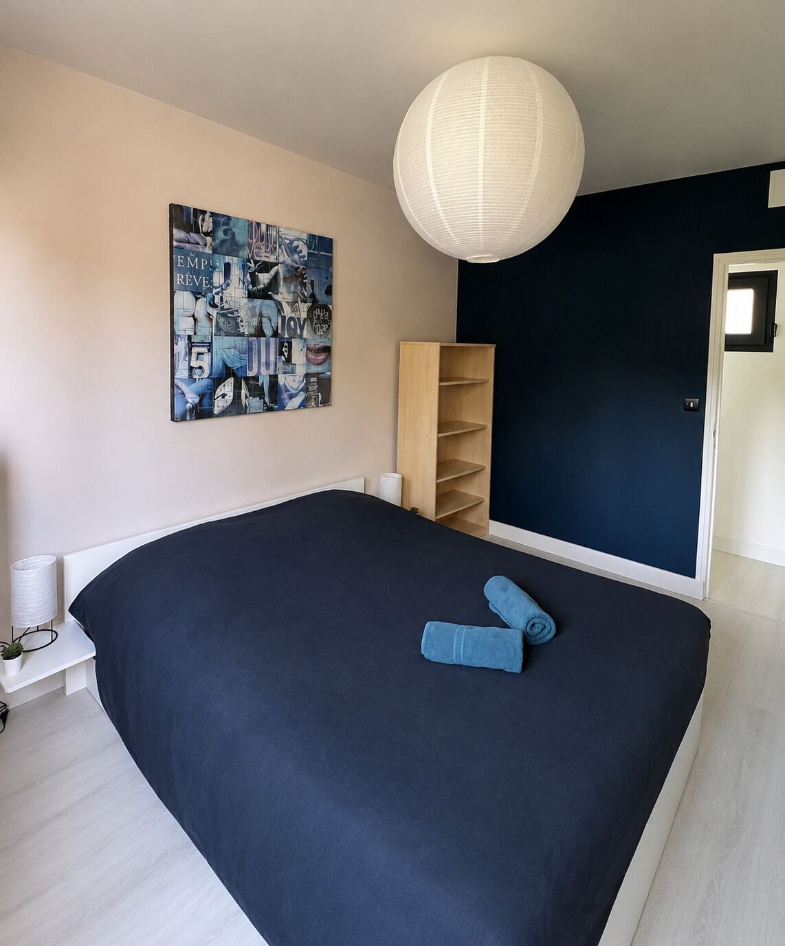 A bedroom with a large bed, a wall-mounted light fixture, a bookshelf, and a poster on the wall.