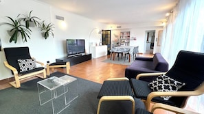 A modern living room with a flat-screen TV, a dining area with a table and chairs, and a kitchen in the background.