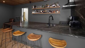Una zona de bar con mostrador de mármol, taburetes de madera, un grifo negro y estantes con botellas de licores.