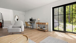 Una sala de estar moderna con suelo de madera, un sillón gris, una mesa de centro de madera, un escritorio con una computadora y un ventanal con vistas a un bosque.