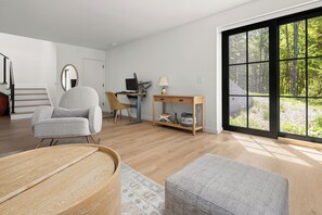 Ein modernes Wohnzimmer mit Holzboden, einem grauen Sessel, einem Couchtisch aus Holz, einem Schreibtisch mit Computer und einem großen Fenster mit Blick auf einen Wald.