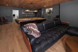 A modern living room with a pool table, leather sofa, wooden flooring, and a bar area with shelves of bottles.