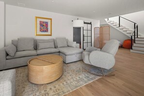 A modern living room with a grey sectional sofa, a round wooden coffee table, a grey armchair, and a staircase.