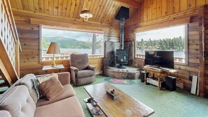 L’intérieur d’une cabane en rondins, avec un canapé, un fauteuil, une télévision, une cheminée et de grandes fenêtres donnant sur une zone boisée.