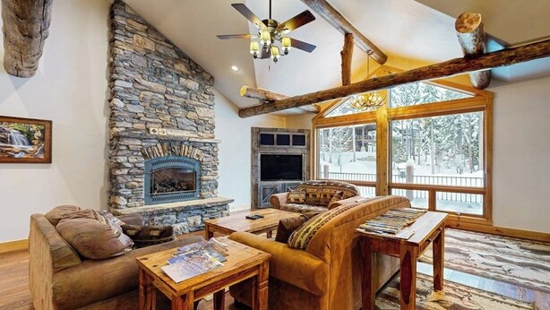 A cozy living room with a stone fireplace, wooden furniture, and large windows.
