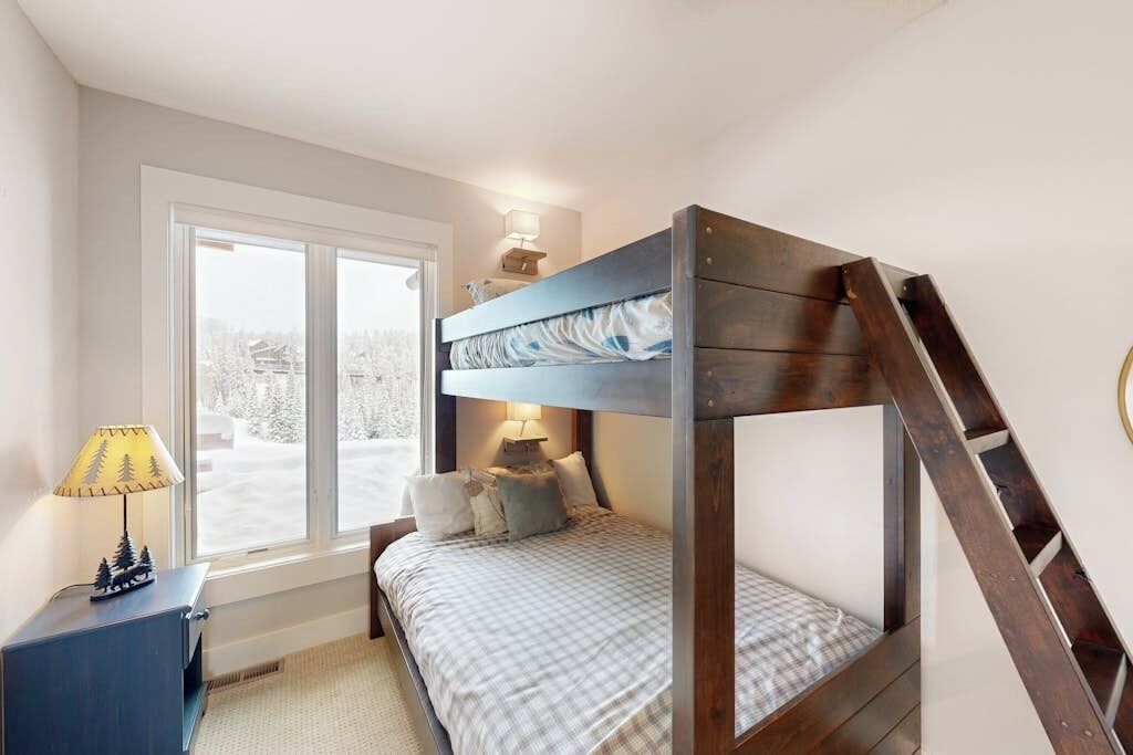 A bedroom with a bunk bed, a desk with a lamp, a window with a view of trees, and a staircase.