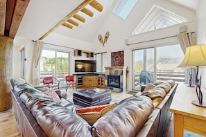 Ein großzügiger Wohnbereich mit Ledersofa, Holztisch und Kamin. Es gibt eine Küchenzeile mit Fernseher und Schränken, sowie große Fenster mit Blick ins Freie.