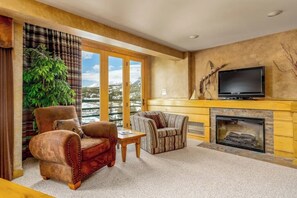 A living room with a fireplace, a television, and a large window with a view of mountains. - Mountain-view condo with heated hot tub, pool, sauna & easy access to slopes (Big Sky)