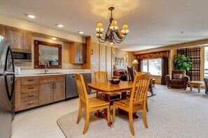 A kitchen with wooden cabinets, a dining table with chairs, a chandelier, and a large window with curtains. - Mountain-view condo with heated hot tub, pool, sauna & easy access to slopes (Big Sky)