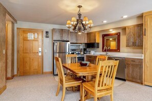 A kitchen with wooden cabinets, a stainless steel refrigerator, a microwave, and a dining table with chairs. - Mountain-view condo with heated hot tub, pool, sauna & easy access to slopes (Big Sky)