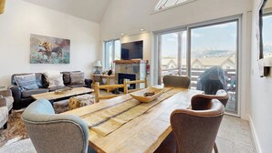 A spacious living room with a large wooden dining table, comfortable seating, a fireplace, and large windows offering a view of the mountains. - Slopeside townhome with ski-in/ski-out and private hot tub (Big Sky)