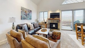 A spacious living room with a fireplace, a large TV, and a view of the outdoors. - Slopeside townhome with ski-in/ski-out and private hot tub (Big Sky)