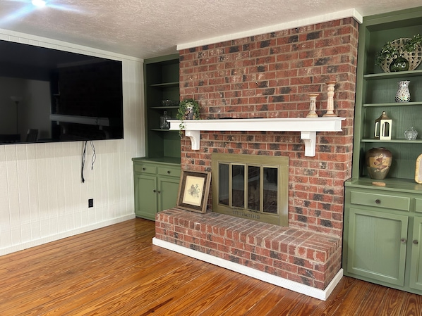 A living room with a brick fireplace, green cabinets, and a mounted television.
