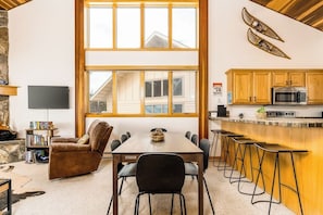 A dining area with a large window, a TV, a bookshelf, and a kitchen with wooden cabinets and a microwave. - Ski-in/ski-out townhome steps from Bear Back lift w/free WiFi & wood fireplace (Big Sky)