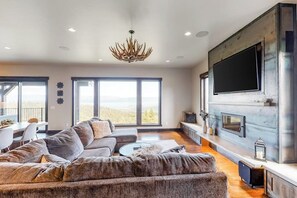 A modern living room with a large flat-screen TV, a fireplace, and a view of the outdoors. - Gorgeous lake-view cabin with game room & fireplaces - near Blacktail Mountain (Lakeside)