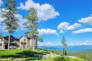 A large wooden house with a garage, surrounded by a grassy lawn and trees, overlooking a mountainous landscape. - Gorgeous lake-view cabin with game room & fireplaces - near Blacktail Mountain (Lakeside)