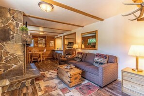 Ein gemütliches Wohnzimmer mit Kamin aus Stein, Holzbalken, einer Couch mit gemusterten Kissen und einem rustikalen Couchtisch.