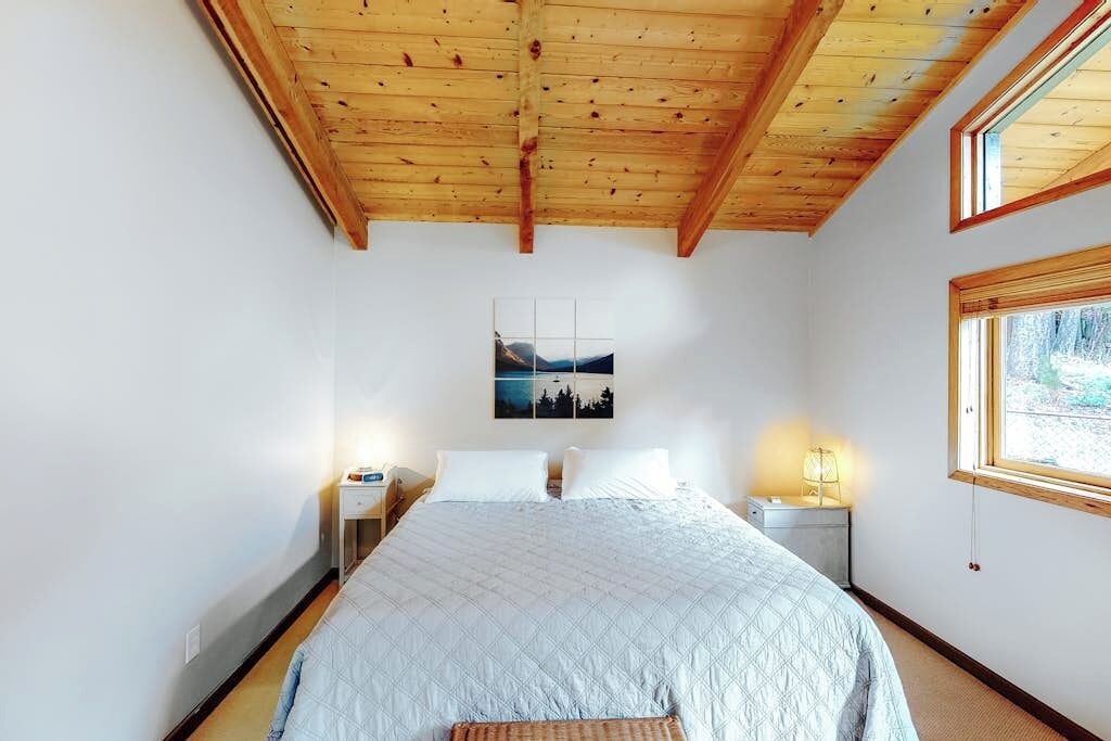 A bedroom with a wooden ceiling, a bed with a quilted bedspread, two bedside tables with lamps, a wall-mounted picture, and a window with a view.