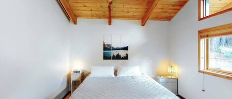A bedroom with a wooden ceiling, a bed with a quilted bedspread, two bedside tables with lamps, a wall-mounted picture, and a window with a view.