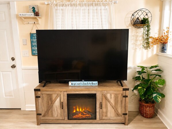 Ein Fernseher auf einer Holzkommode, darunter ein Kamin, rechts eine Pflanze und ein Schild an der Wand.