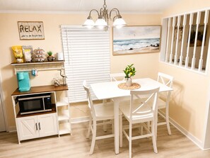 A dining area with a white table and chairs, a kitchen with a microwave and cabinets, a window with blinds, and a beach-themed painting. - Home Away from Home in Little River (Little River)