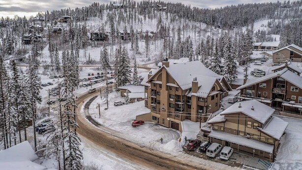 Un edificio de varios pisos con un paisaje de estación de esquí al fondo, rodeado de árboles cubiertos de nieve.