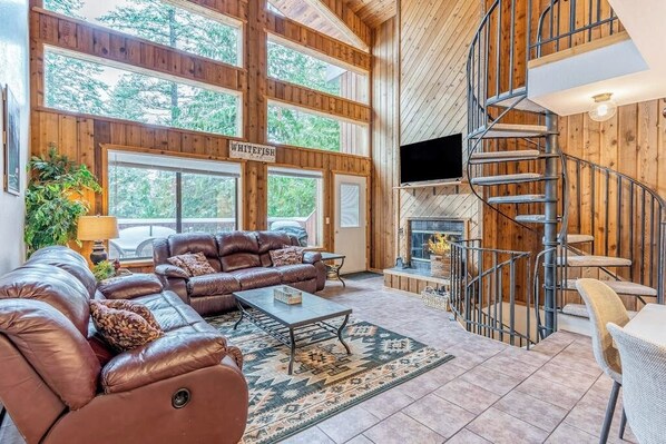 Ein geräumiges Wohnzimmer mit einem großen braunen Ledersofa, einem Couchtisch und einem Fernseher. Eine Holztreppe und ein Schild mit der Aufschrift 'WHITEFISH' sind ebenfalls vorhanden.