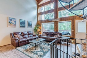 Ein geräumiges Wohnzimmer mit zwei Ledersofas, einem Couchtisch und einem gemusterten Teppich. Große Fenster und eine Treppe, die in einen oberen Bereich führen.