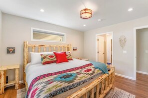 A bedroom with a wooden bed frame, a colorful bedspread, a bedside table, a nightstand, a ceiling light fixture, and a door leading to another room. - Relaxing single-level home with a patio, washer/dryer, & central AC (Lakeside)