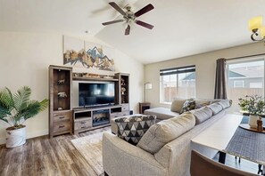 A living room with a flat-screen TV, a ceiling fan, a sofa, a dining table, and a large window. - Bridger View Retreat | House in Belgrade with enclosed backyard, patio, & grill (Belgrade)
