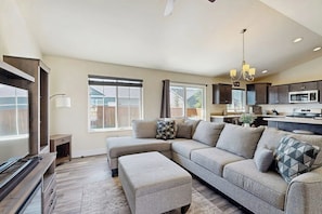 A modern living room with a sectional sofa, a TV, and a kitchen in the background. - Bridger View Retreat | House in Belgrade with enclosed backyard, patio, & grill (Belgrade)