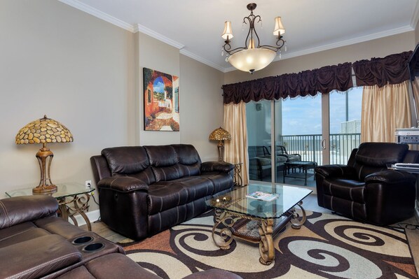 A living room with a leather sofa, glass coffee table, and a chandelier. - Beachfront Fun in the heart of Biloxi! Pools, sunsets, and steps to the sand! (Biloxi)