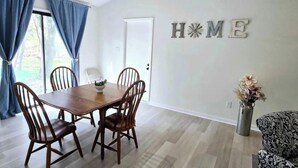 Ein Essbereich mit Holztisch und Stühlen, einem 'HOME'-Schild an der Wand und einem gemusterten Sofa.