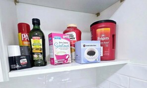 En hylde med forskellige køkkenartikler, heriblandt en flaske olivenolie, en sødekasse, en beholder med kaffefiltre og en dåse Folgers kaffe.