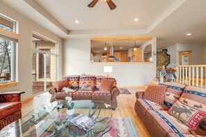 A spacious living room with a glass coffee table, two sofas with patterned cushions, a wooden ceiling fan, and a view of the kitchen through an open doorway. - Spacious 2-story home with deck, fireplace, & W/D - near Glacier National Park (Whitefish)