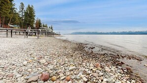 A pebbled beach with a wooden pier extending into calm water, surrounded by trees. - Lakefront home with amazing mountain views, wraparound deck, fireplace, & grill (Lakeside)