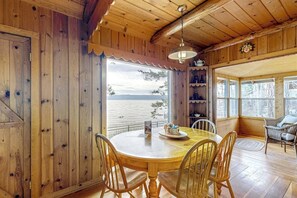 A wooden cabin with a round dining table and chairs, a view of the water, and a cozy interior with shelves and a hanging light. - Lakefront home with amazing mountain views, wraparound deck, fireplace, & grill (Lakeside)