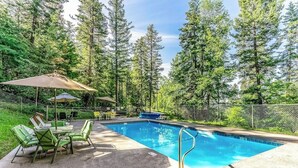 An outdoor swimming pool with a metal ladder, surrounded by a fence, and green chairs and tables under a canopy. - Charming condo with lake access, shared pools and hot tub, WiFi, and fireplace (Whitefish)