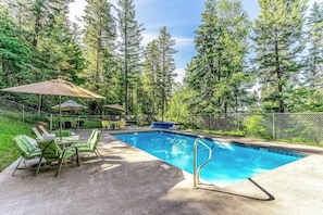 An outdoor swimming pool with a metal ladder, surrounded by a fence, and green chairs and tables under a canopy. - Charming condo with lake access, shared pools and hot tub, WiFi, and fireplace (Whitefish)