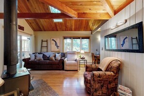 A cozy living room with a fireplace, wooden ceiling beams, a sofa, a chair, and artwork on the walls. - Cozy condo with deck, wood stove, pool, sauna, tennis - near skiing & fishing (Whitefish)