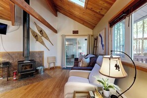 A cozy living room with a fireplace, a sofa, a lamp, and a view of the outdoors. - Cozy condo with deck, wood stove, pool, sauna, tennis - near skiing & fishing (Whitefish)