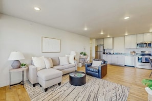A modern living room with a sofa, armchair, and coffee table, connected to a kitchen with white cabinets and appliances.