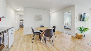 Een moderne keuken met witte kastjes, een eettafel met stoelen, een aan de muur bevestigde televisie en een plant op de vloer.