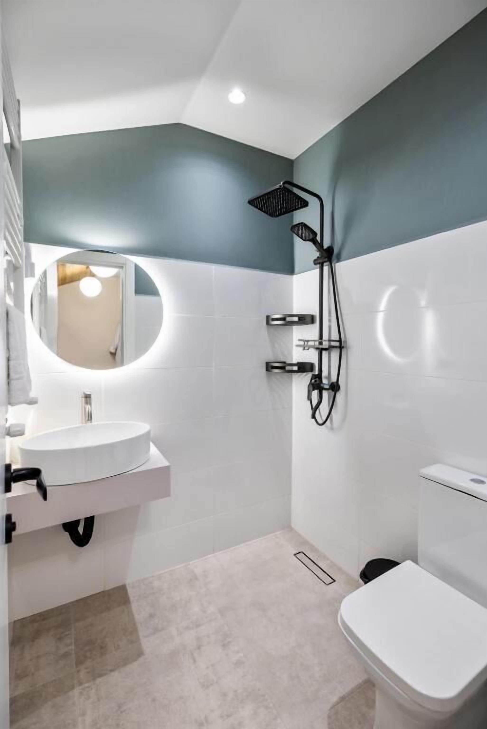 Un bagno moderno con un lavandino bianco, uno specchio rotondo, una doccia con un tubo nero e un water bianco.