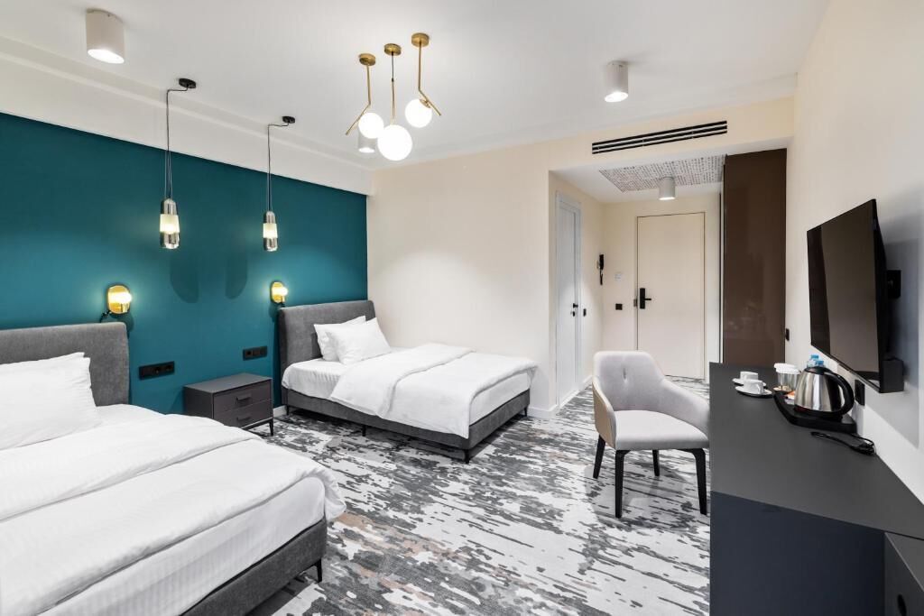 A modern hotel room with a bed, bedside tables, a desk with a chair, a TV, and a unique teal accent wall.