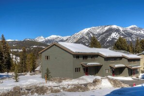 A two-story lodge with a snow-covered roof, situated in a mountainous area with visible peaks in the background. - Townhome minutes from slopes (Big Sky)