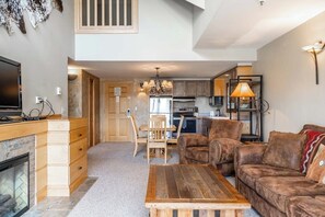 A spacious living area with a fireplace, wooden furniture, and a kitchen in the background. - Slope side family fun condo with shared pool and hot tub (Big Sky)